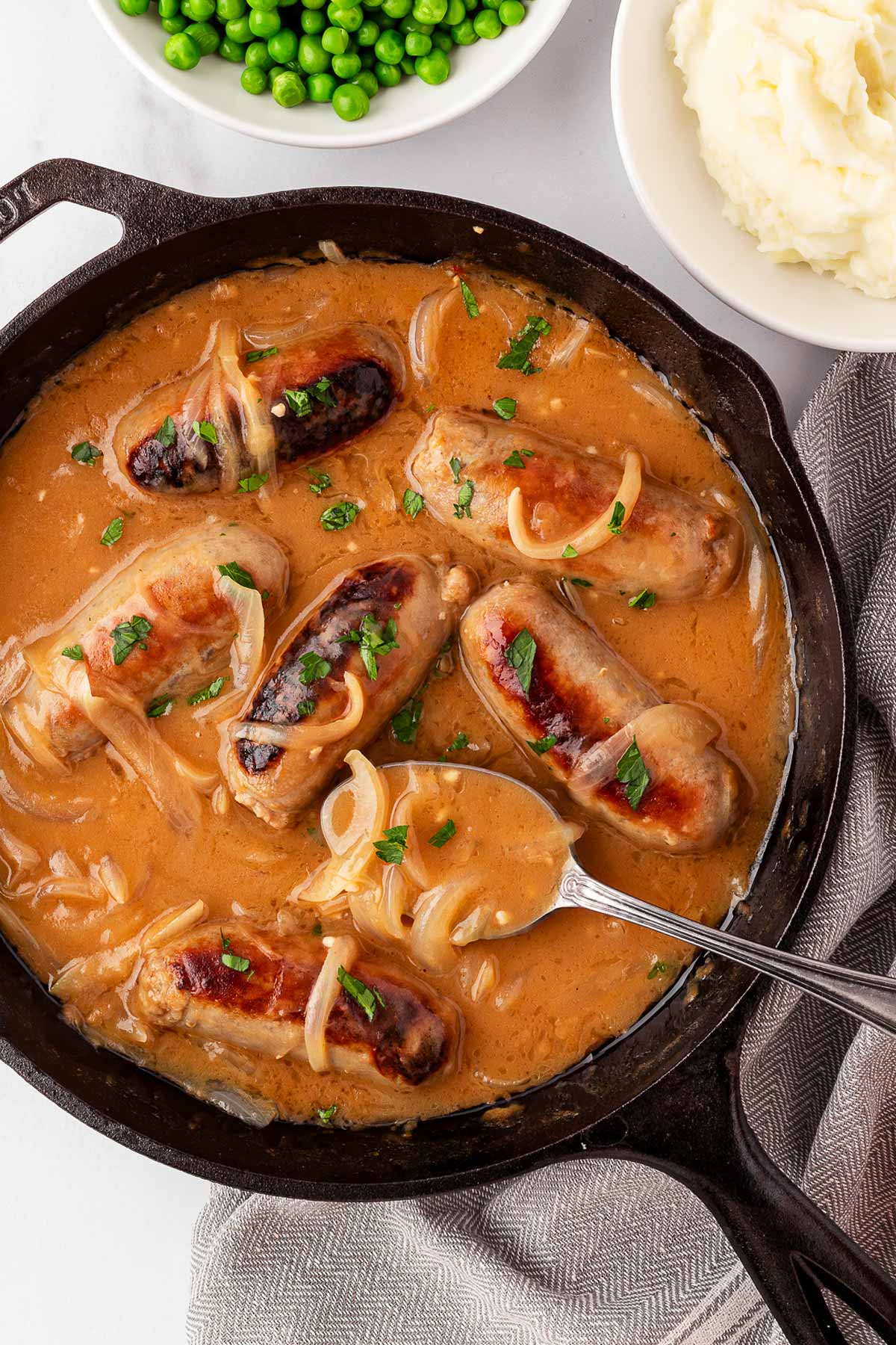 overhead view of bangers and mash in cast iron skillet with bowls of mash and peas