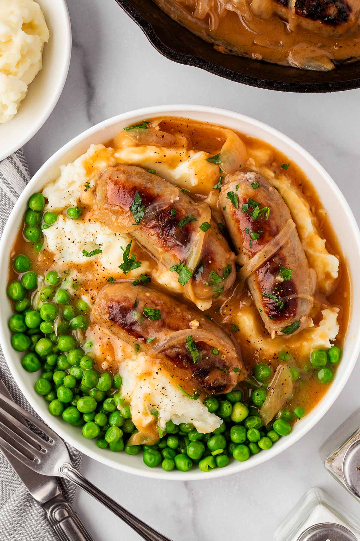 overhead view of mashed potatoes, peas and sausages with onion gravy in white bowl