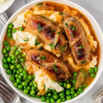 overhead view of mashed potatoes, peas and sausages with onion gravy in white bowl