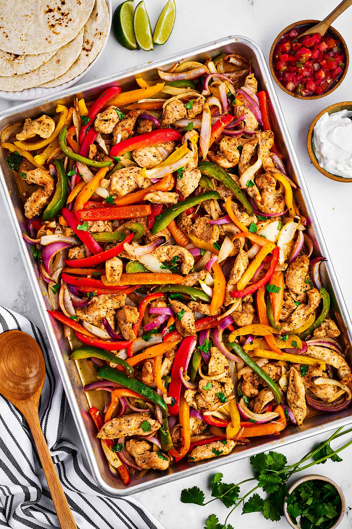 overhead view of baked chicken fajita mixture on sheet pan surrounded by toppings on wheat tortillas