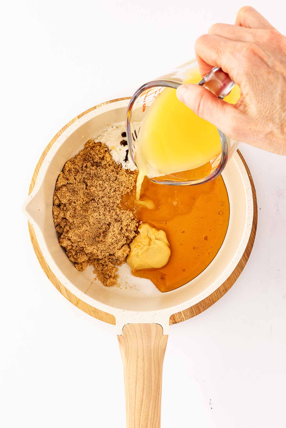 overhead view of female hand pouring orange juice over brown sugar and honey in white saucepan