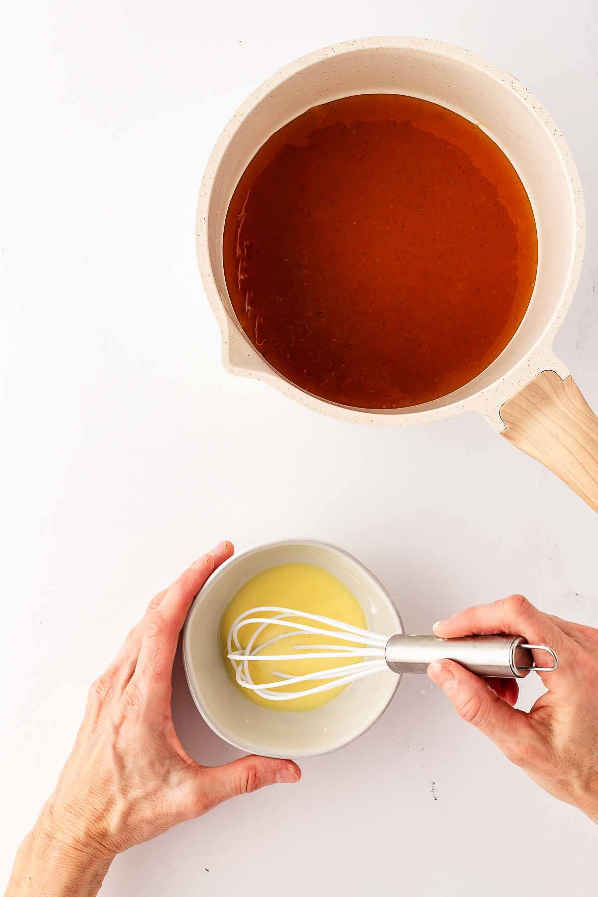 overhead view of female hands whisking cornstarch slurry in small bowl next to saucepan with ham glaze