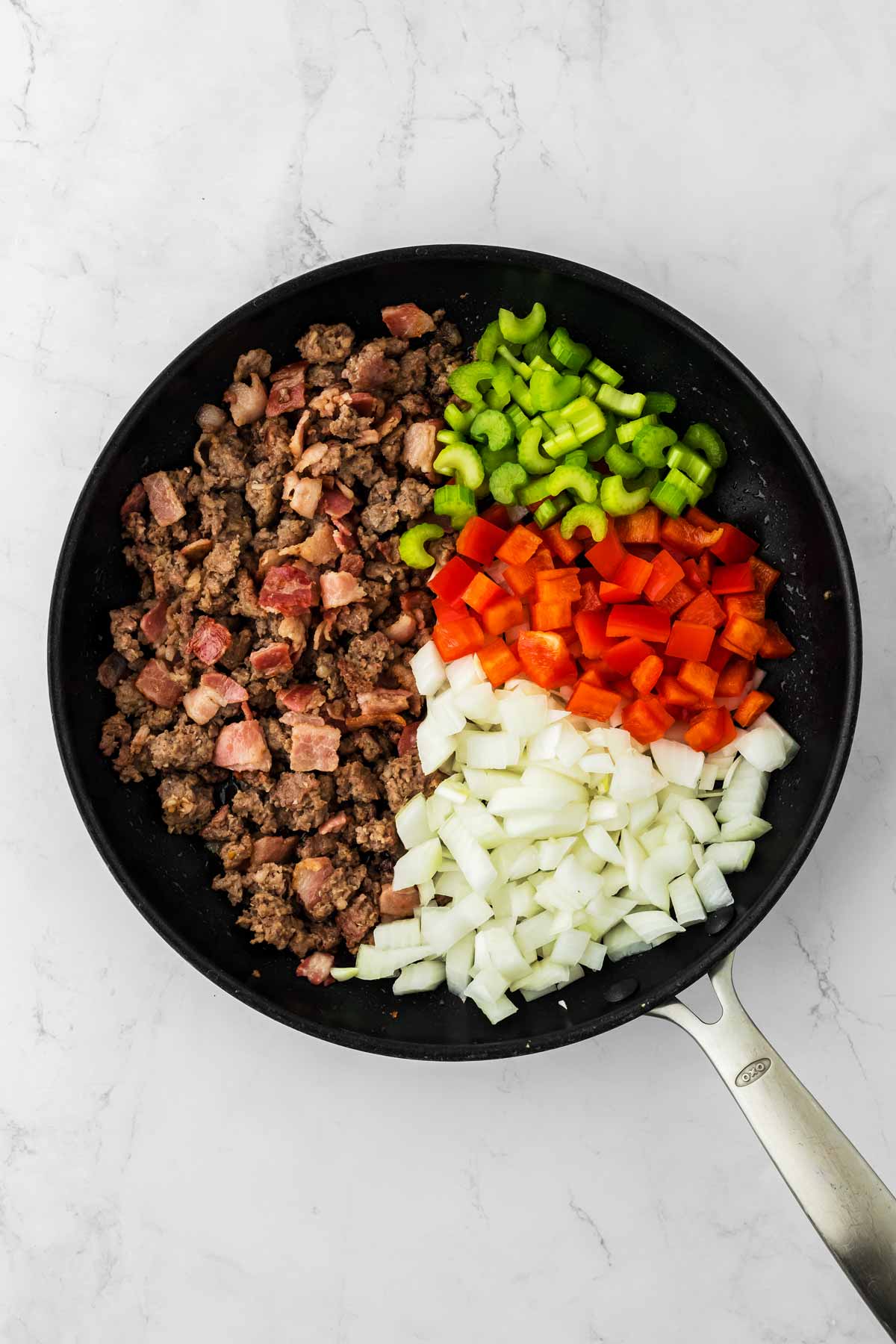 overhead view of cooked sausage and bacon in skillet with raw red pepper, raw celery and raw onion