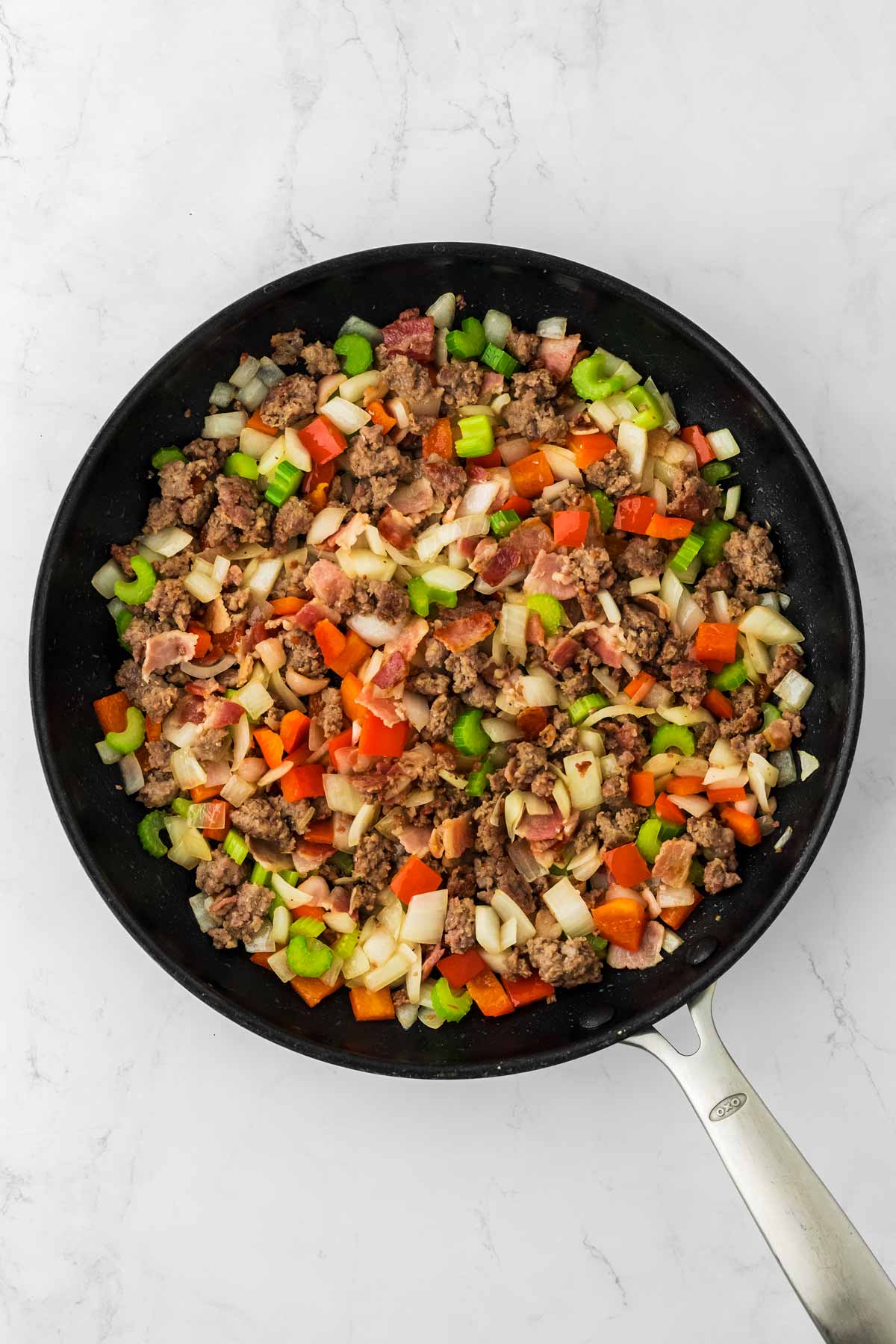 overhead view of cooked sausage and bacon in skillet with cooked red pepper, cooked celery and cooked onion