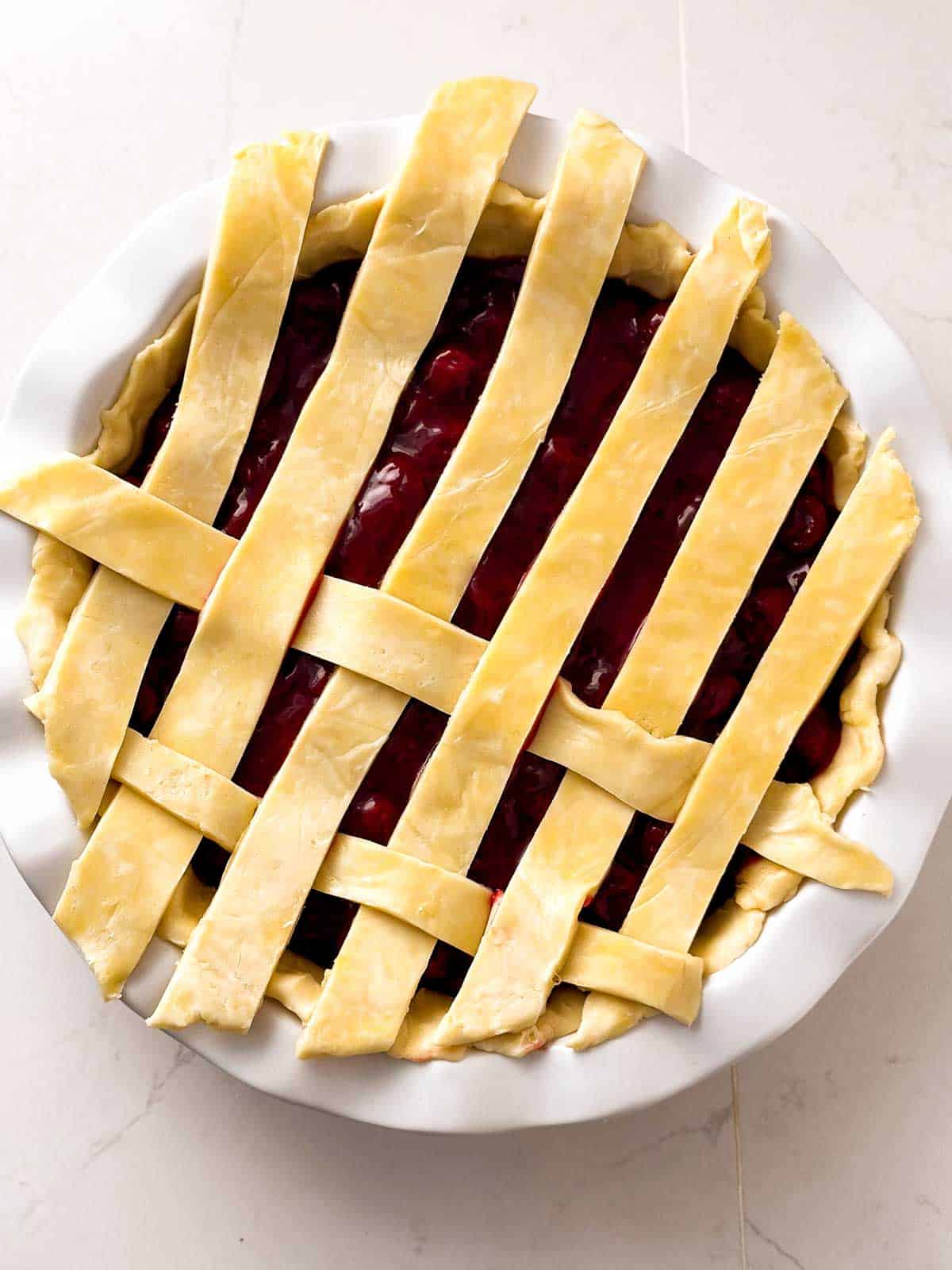 overhead view of pie crust strips on top of unbaked cherry pie assembled in half a lattice weave