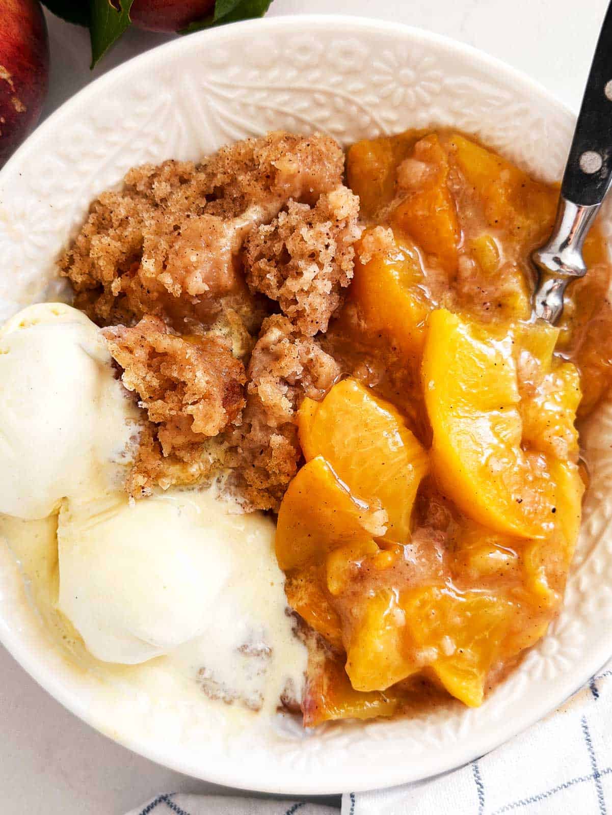 overhead view of crock pot peach cobbler in white bowl with spoon and two scoops of vanilla ice cream