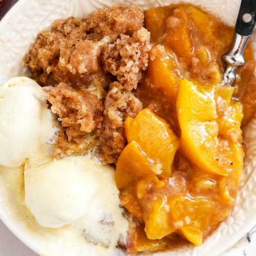 overhead view of crock pot peach cobbler in white bowl with spoon and two scoops of vanilla ice cream