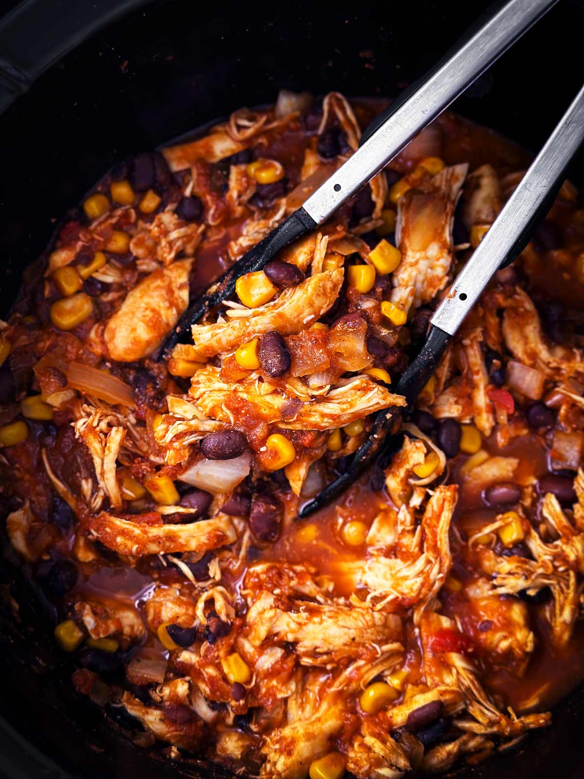 overhead view of crock pot chicken taco mixture in black slow cooker insert with kitchen tongs stuck in