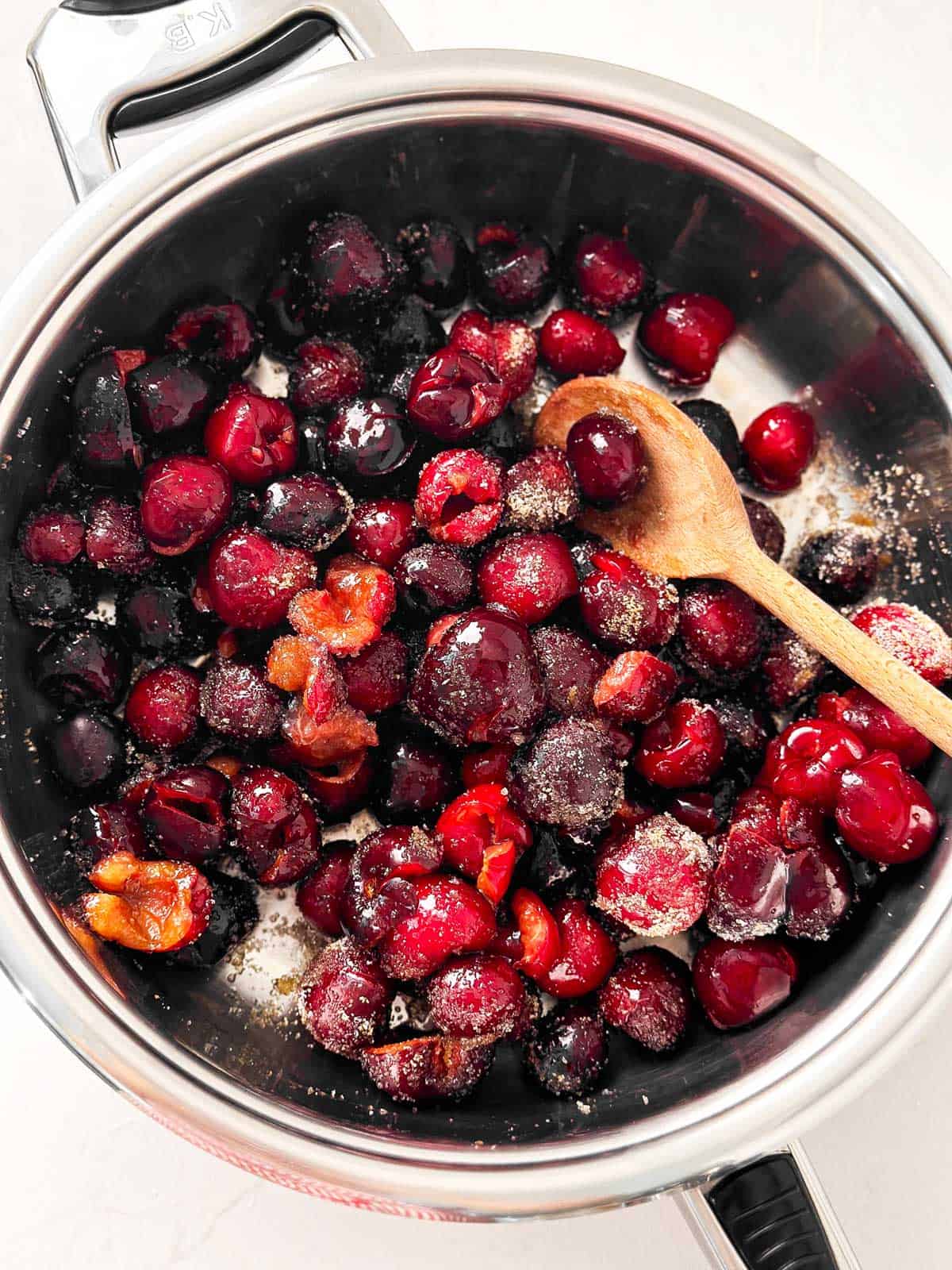 overhead view of uncooked pitted cherries in pan with sugar and wooden spoon