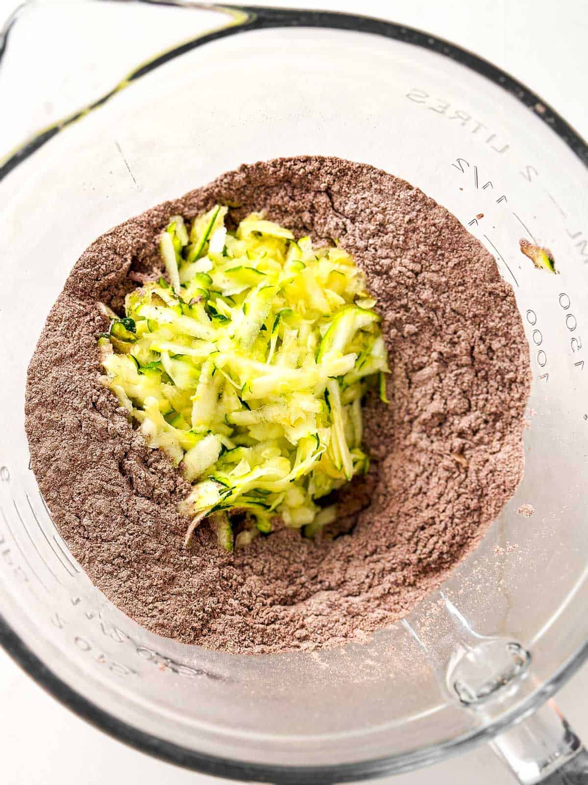 raw shredded zucchini on top of dry ingredients for chocolate zucchini bread in glass bowl