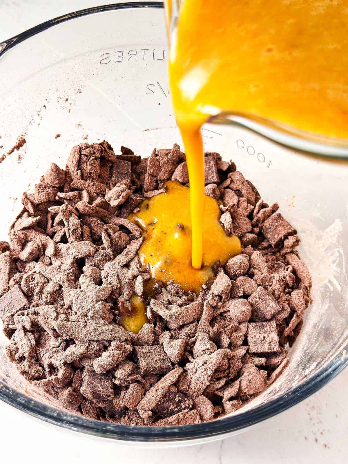 egg mixture pouring from measuring jug over dry chocolate zucchini bread ingredients in glass bowl