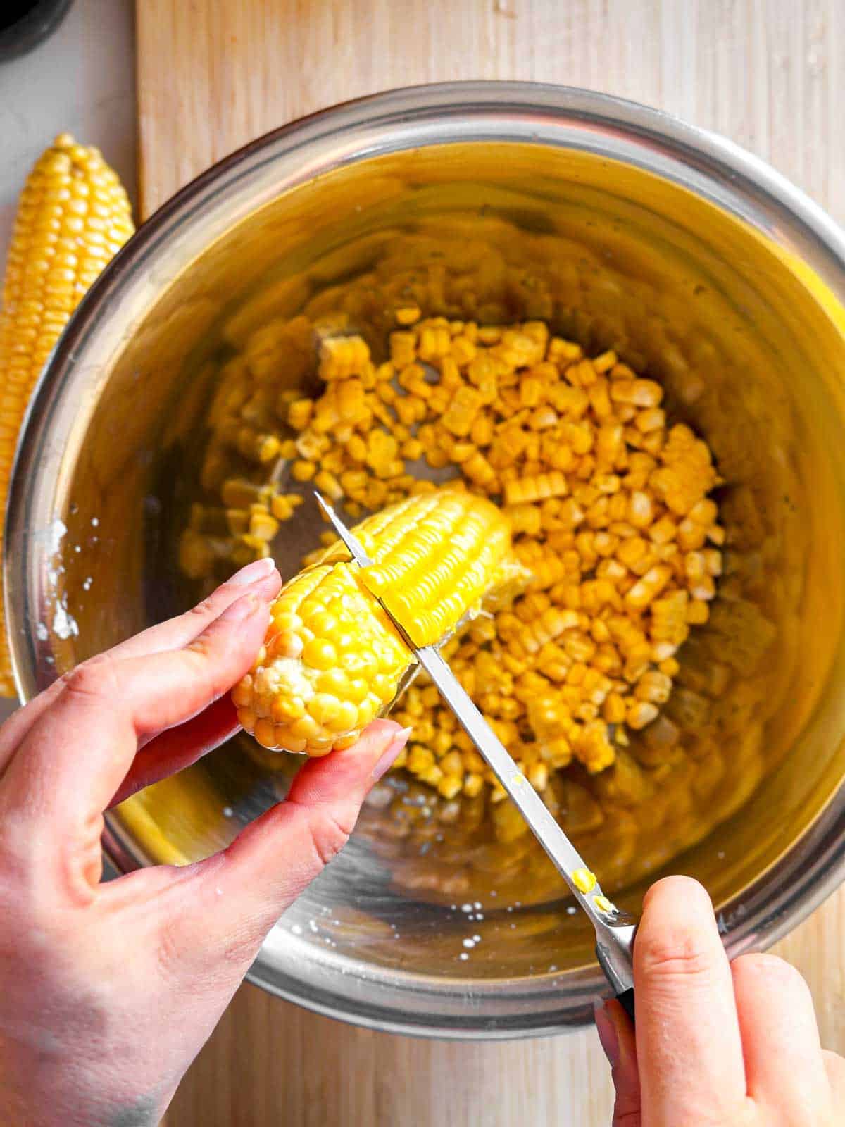 overhead view of female hands cutting kernels off corn ear over metal bowl