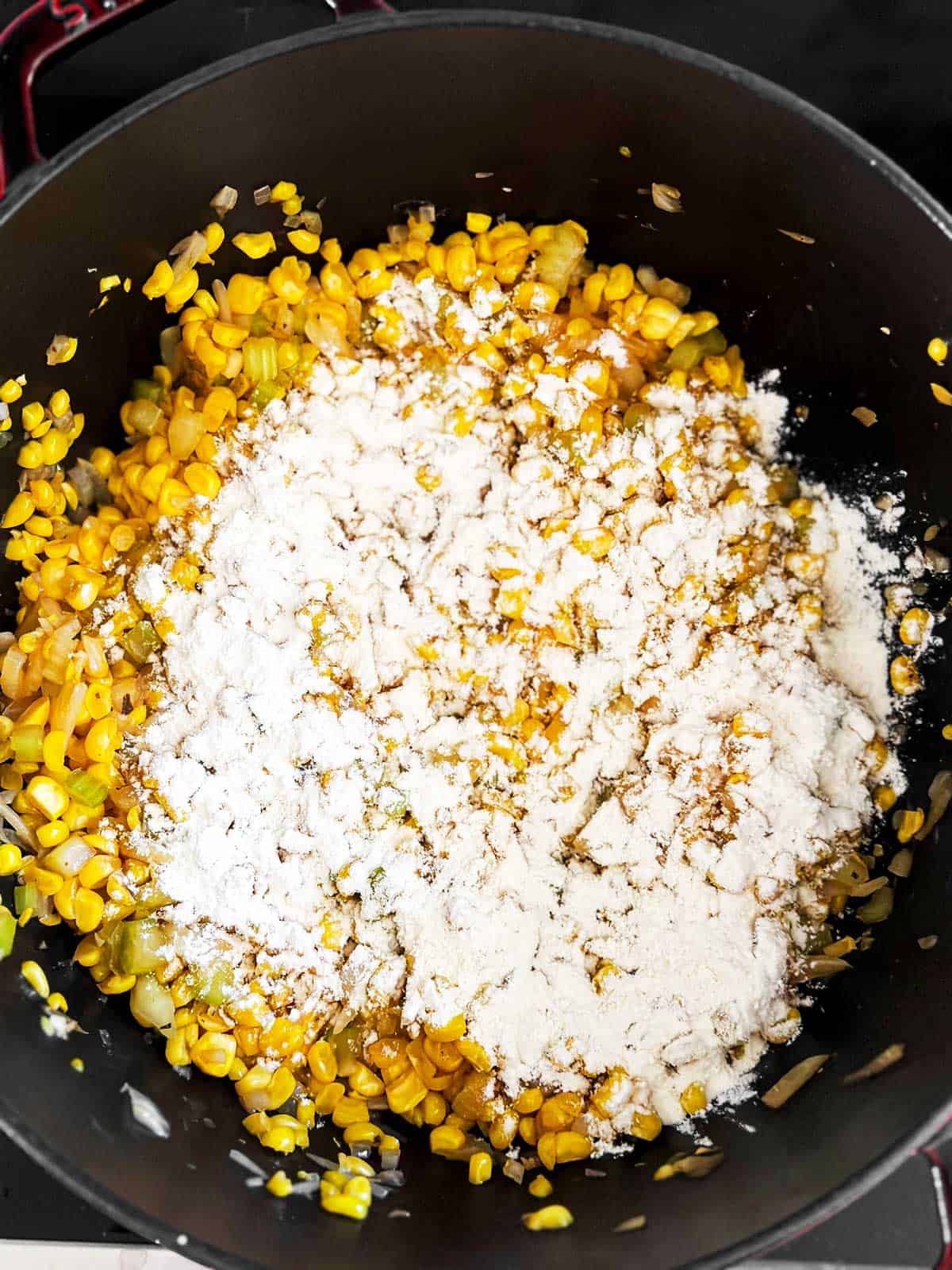 overhead view of flour sprinkled over cooked corn, onion and celery in black Dutch oven