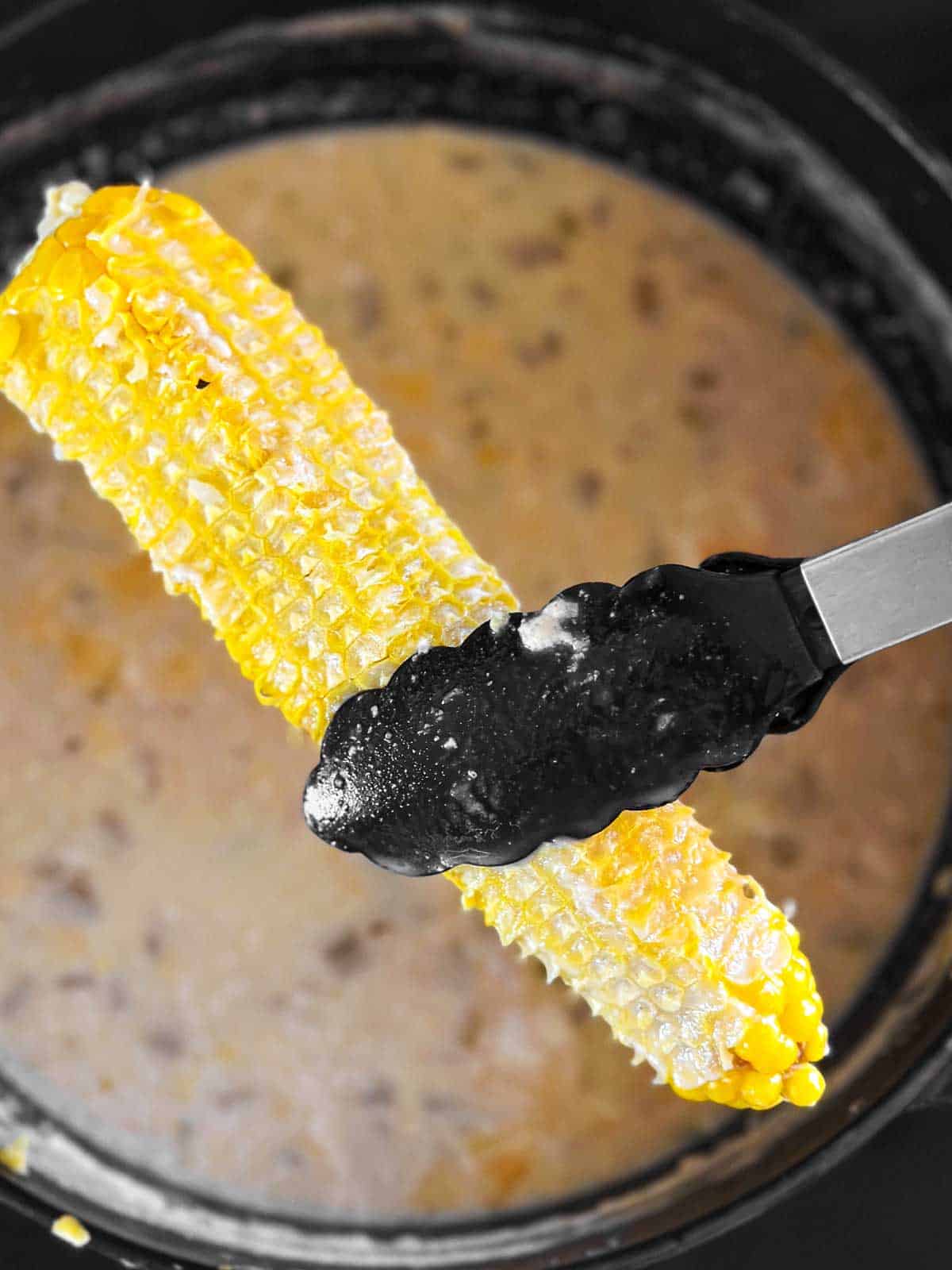 kitchen tongs holding empty corn cob over pot with corn chowder