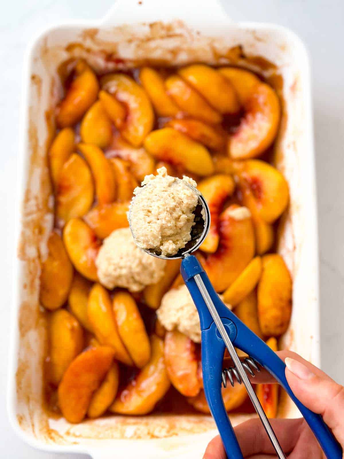 overhead view of female hand holding cookie scoop with dough over peach cobbler filling