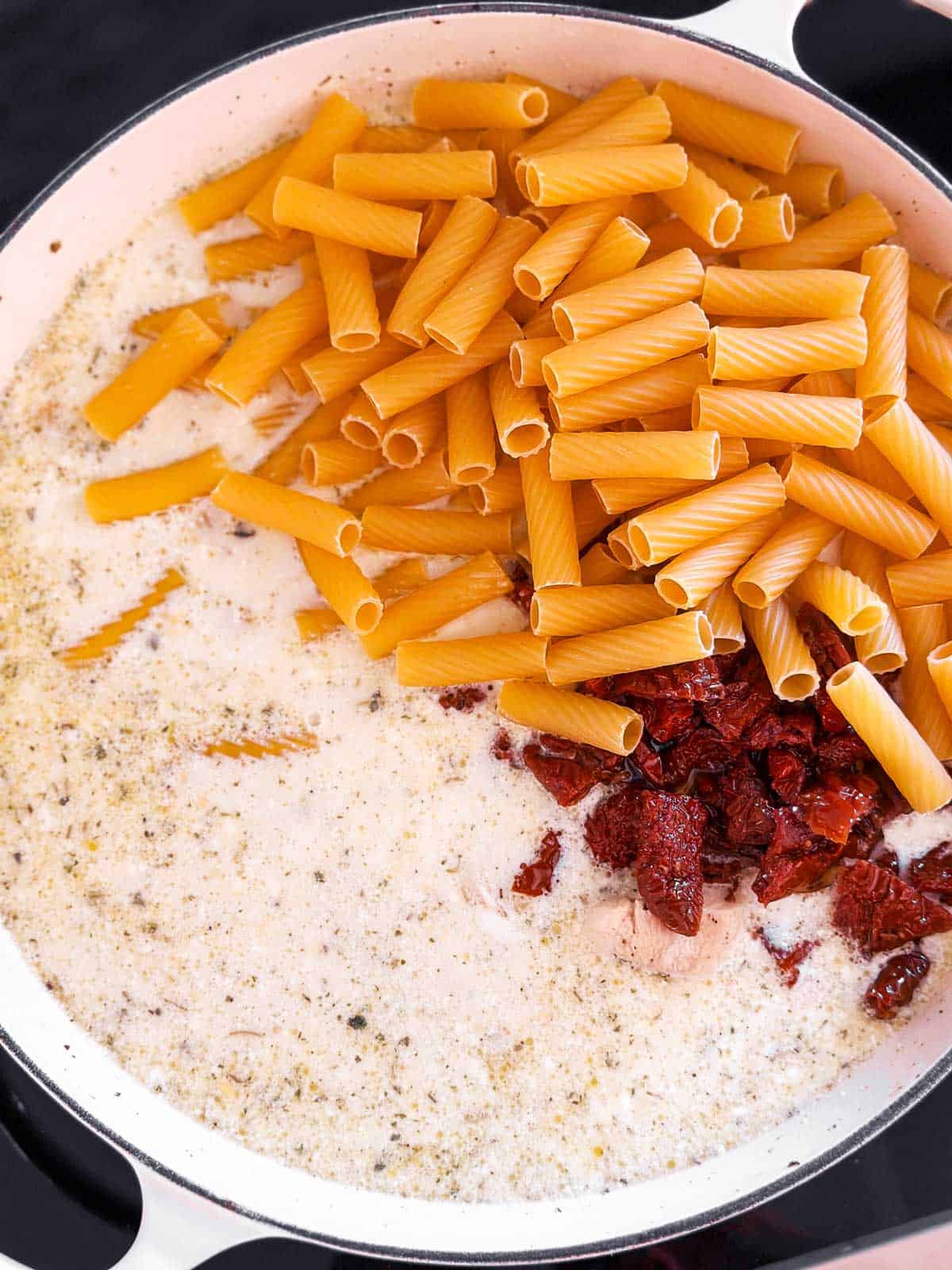 overhead view of sun-dried tomatoes and uncooked pasta in cream sauce in skillet