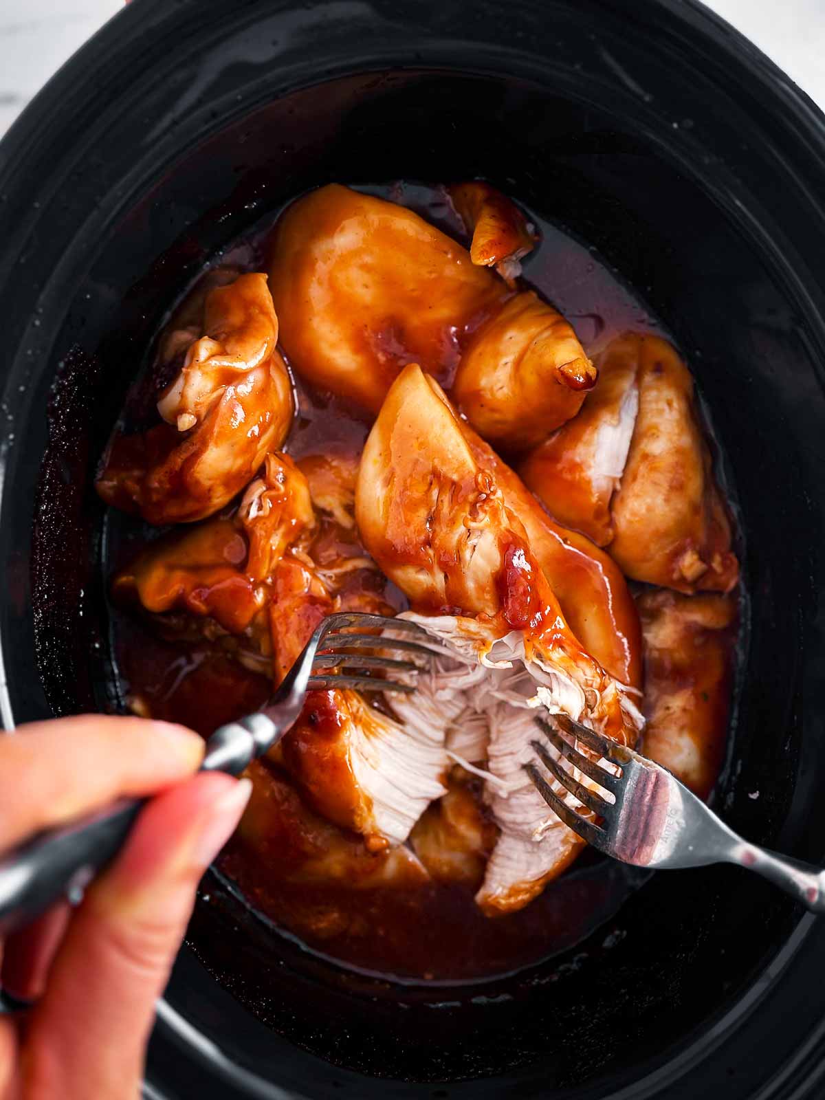 overhead view of female hands shredding chicken in crock using forks