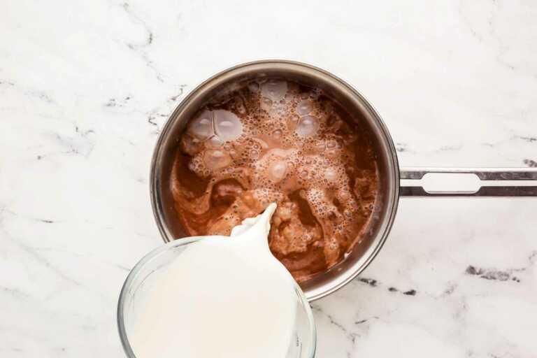 overhead view of milk pouring from glass measuring jug into cocoa milk mix in saucepan