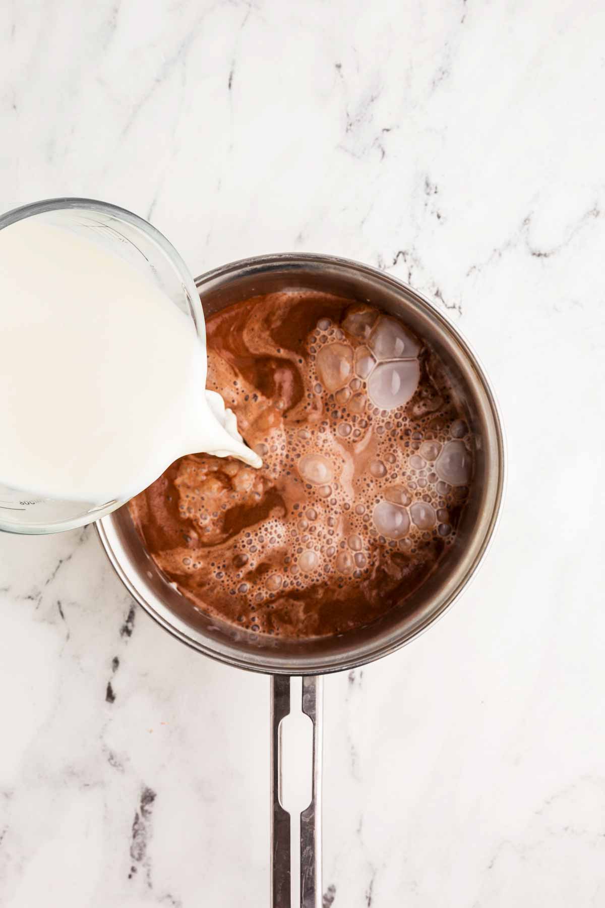 overhead view of milk pouring from glass measuring jug into cocoa milk mix in saucepan