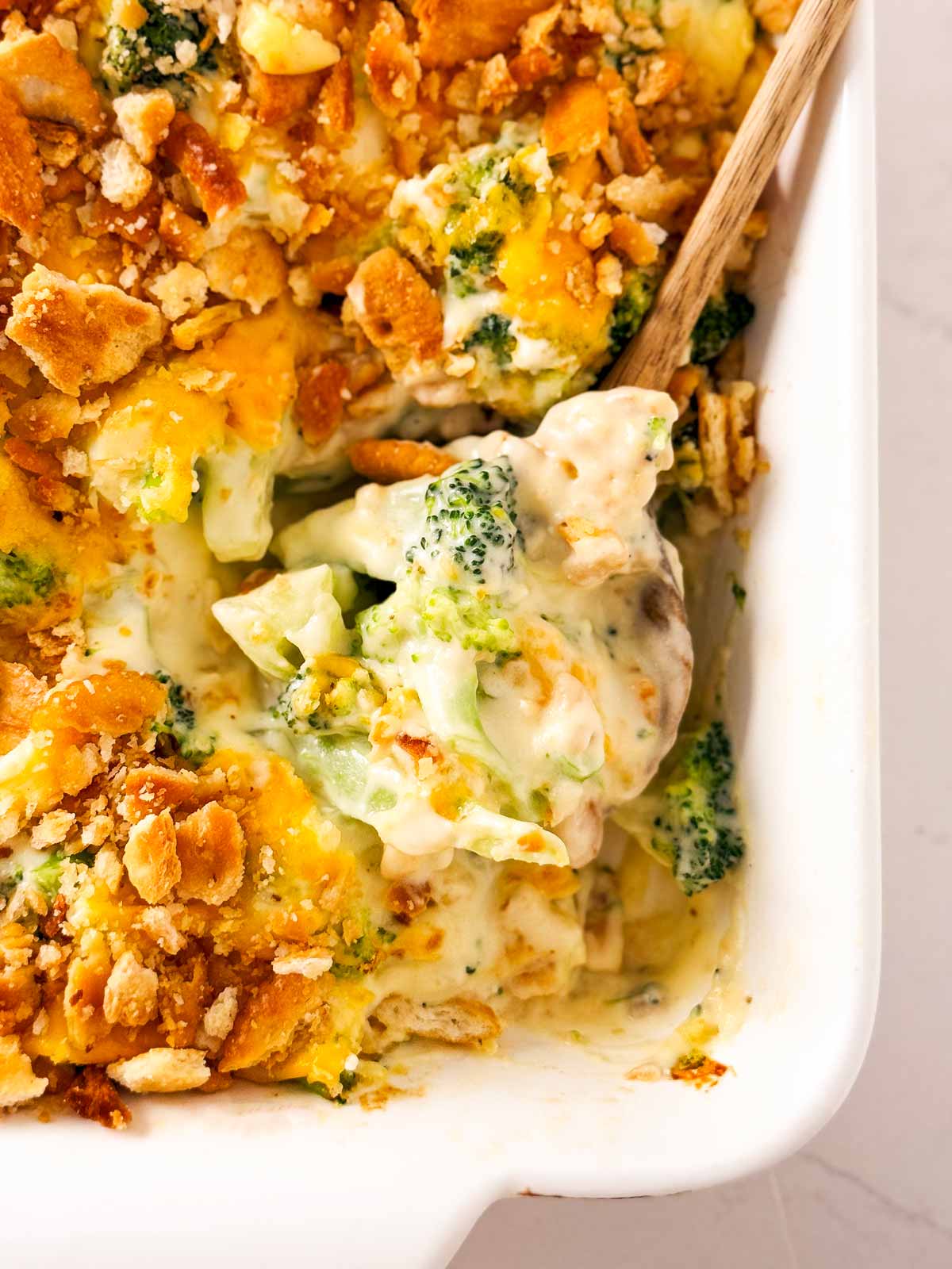 frontal close up view of wooden spoon scooping up broccoli cheese casserole from a white baking dish