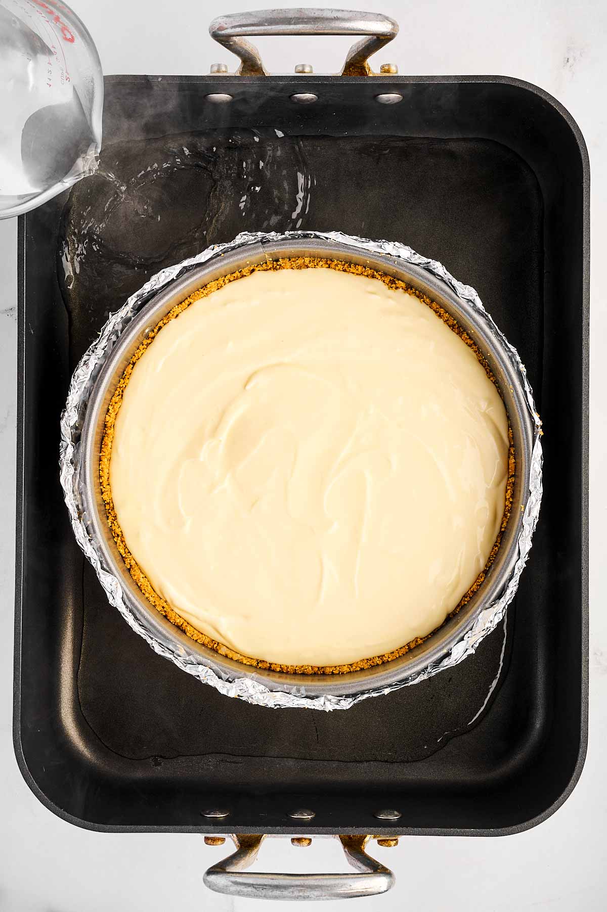 overhead view of springform pan with unbaked cheesecake placed inside roasting pan with hot water being poured into the roasting pan