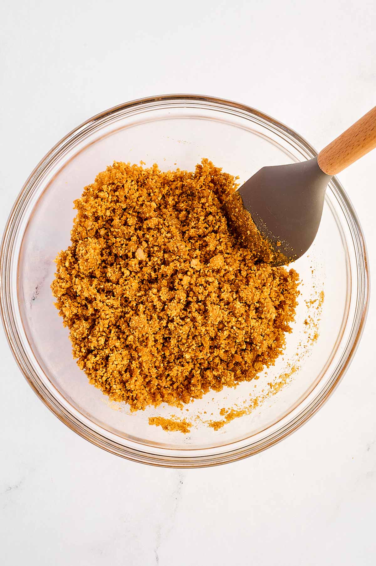 overhead view of graham cracker crumbs, sugar, and melted butter being mixed in a glass bowl with a rubber spatula