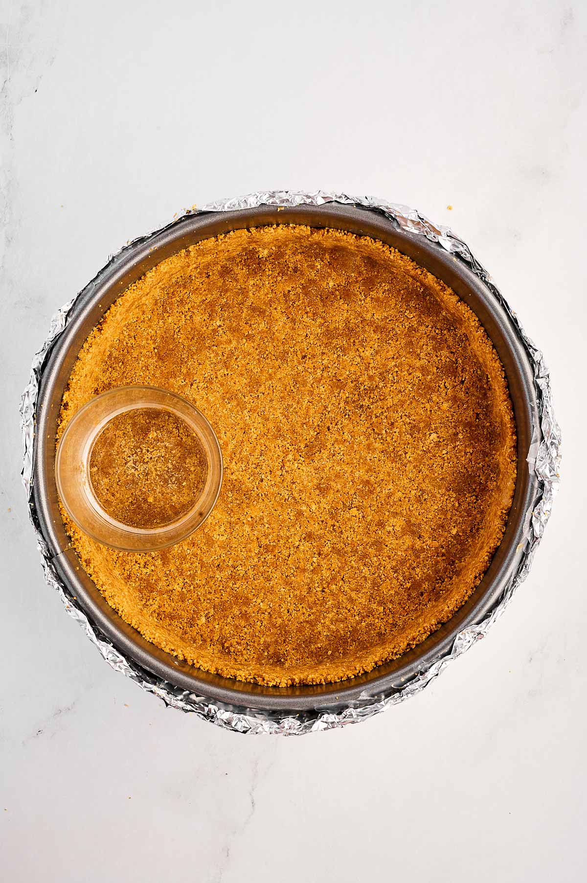 overhead view of graham cracker crust mixture being pressed into the bottom and sides of the springform pan with the back of a glass cup