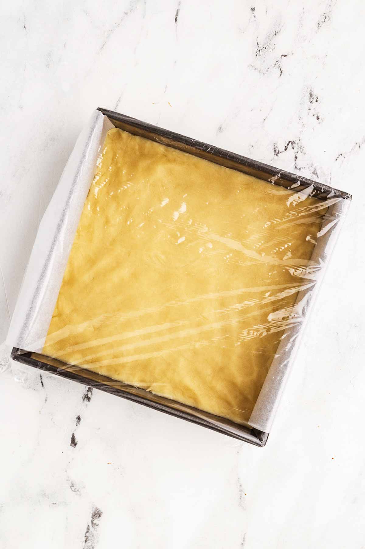 overhead view of lined square baking pan filled with shortbread cookie dough wrapped in plastic wrap