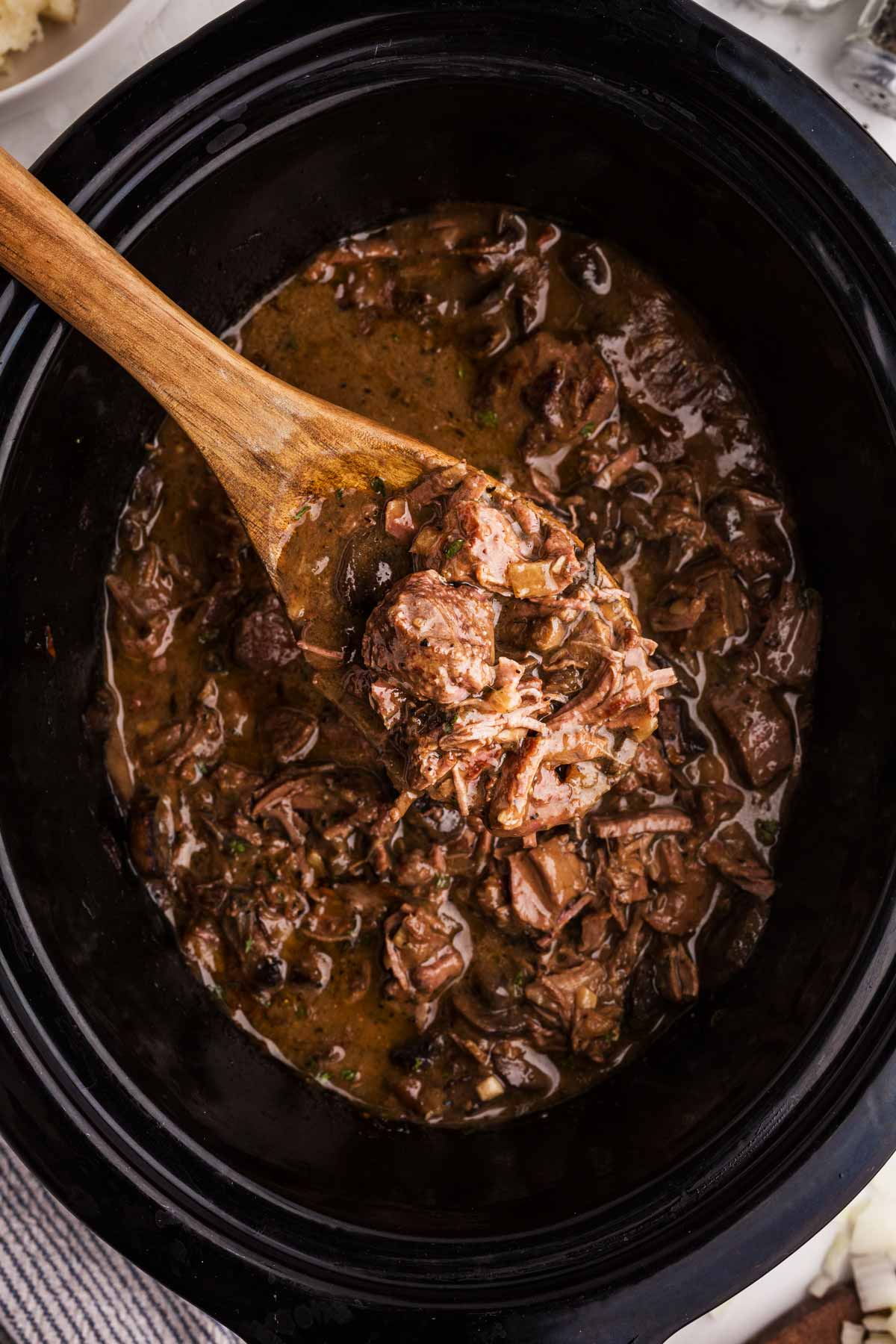 overhead view of wooden spoon scooping beef tips and mushroom gravy from slow cooker