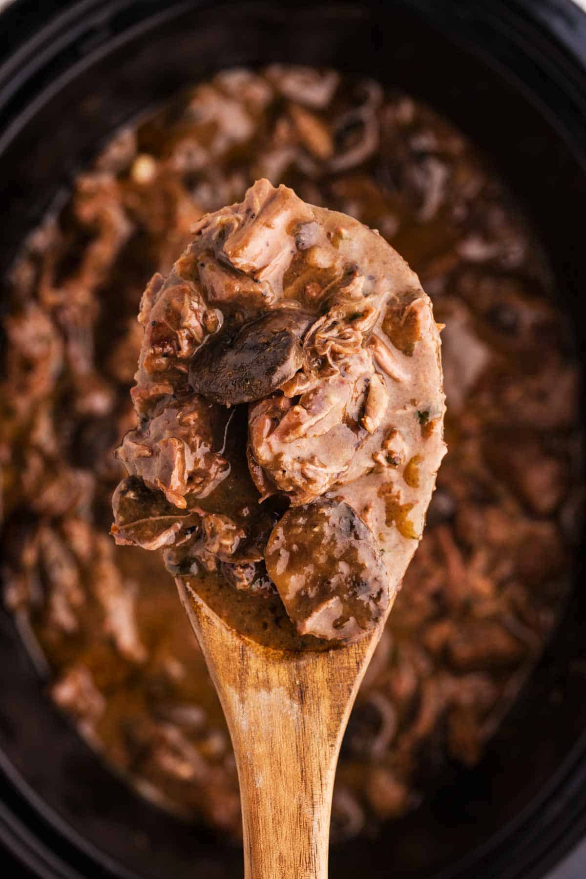 overhead view of beef tips and mushroom gravy on wooden spoon over black crock