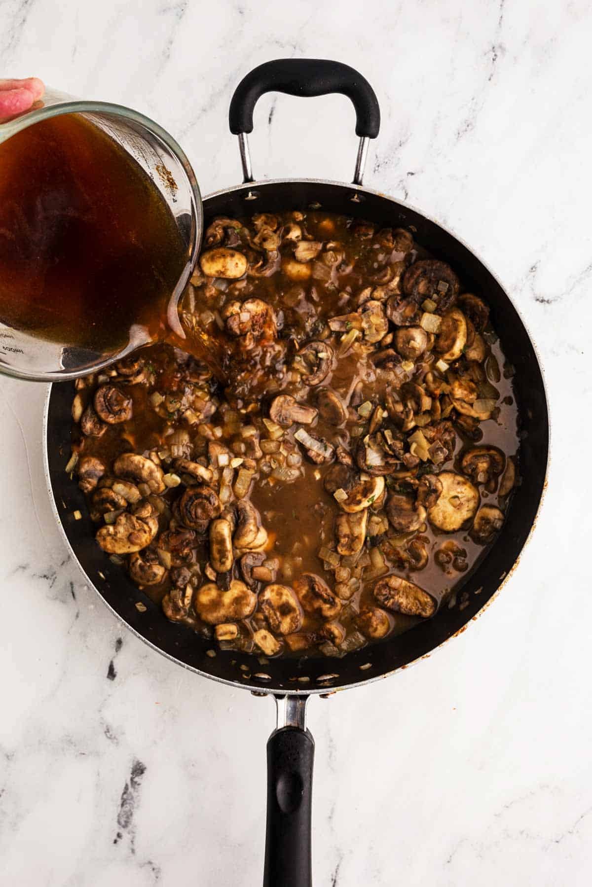 beef broth pouring from glass measuring jug into cooked mushrooms