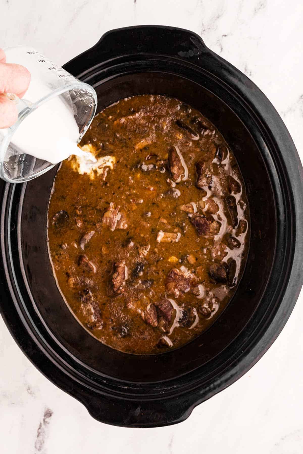 cornstarch slurry pouring from glass measuring jug into beef tips and gravy in slow cooker