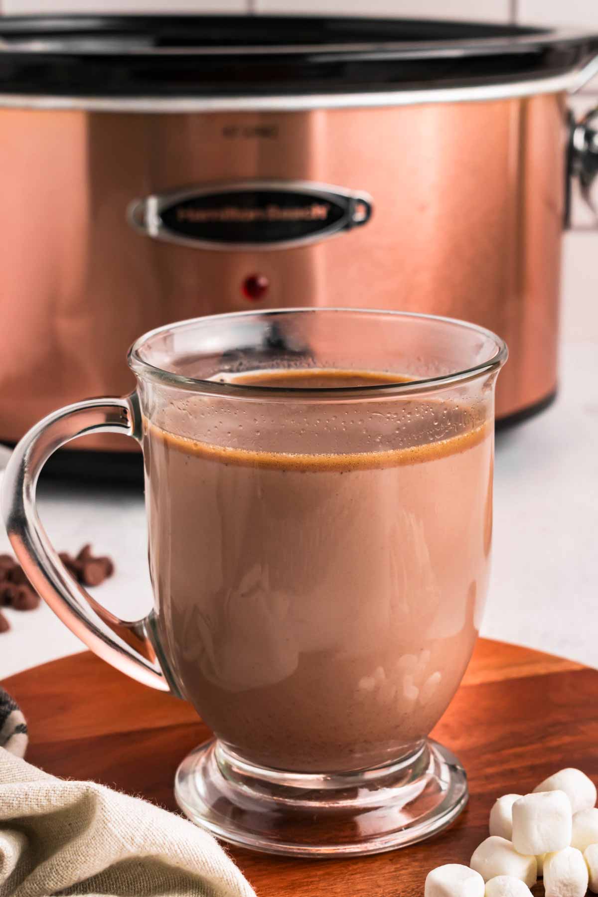 frontal view of glass mug with hot chocolate in front of slow cooker