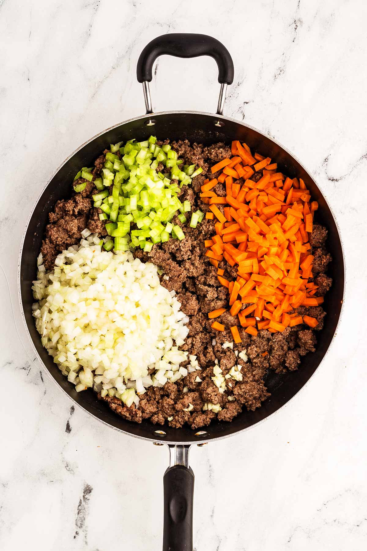 overhead view of raw onions, carrots, celery, and garlic on top of browned ground beef in skillet