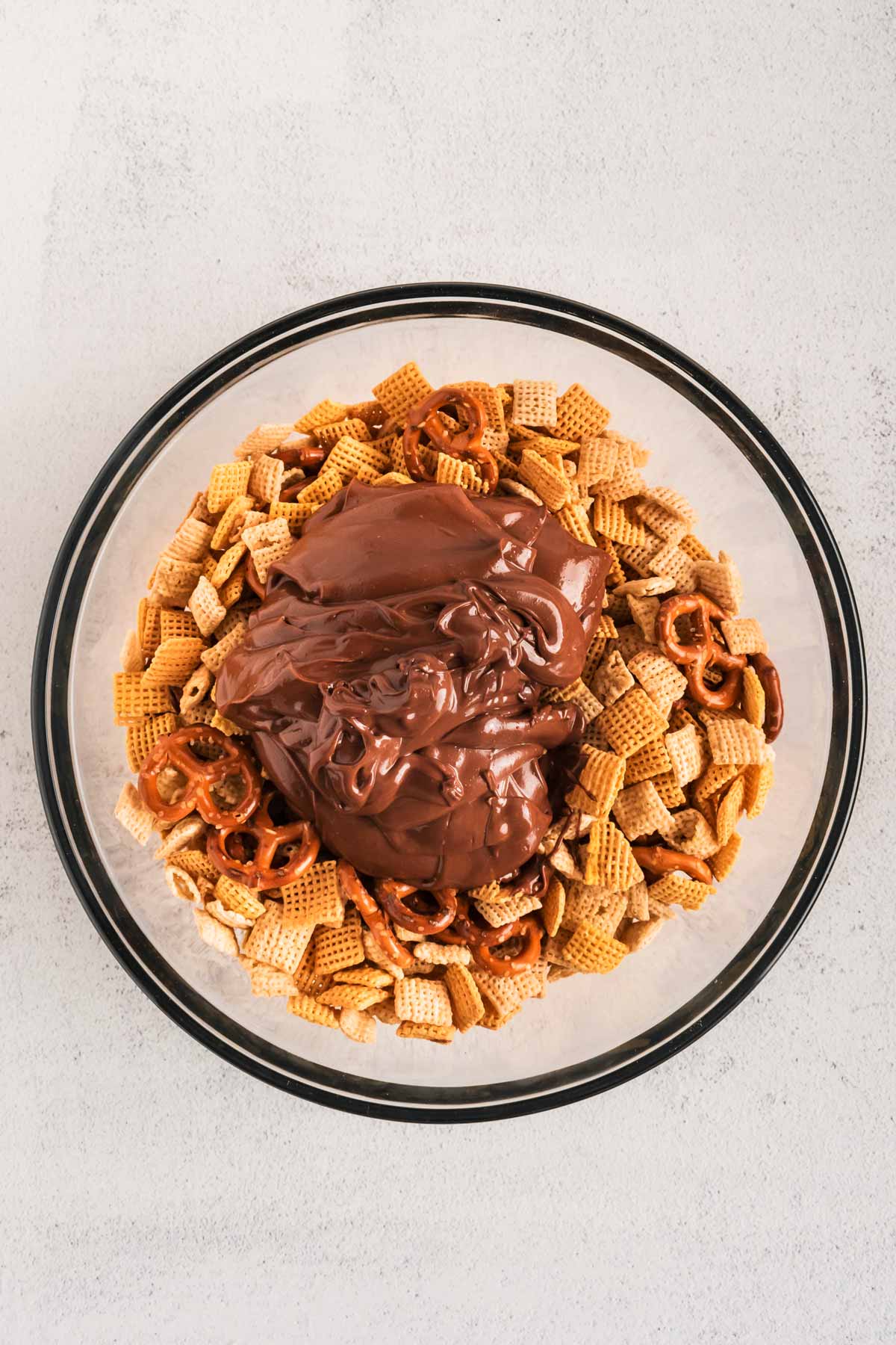 overhead view of melted chocolate on top of cereal and pretzel mix in glass bowl