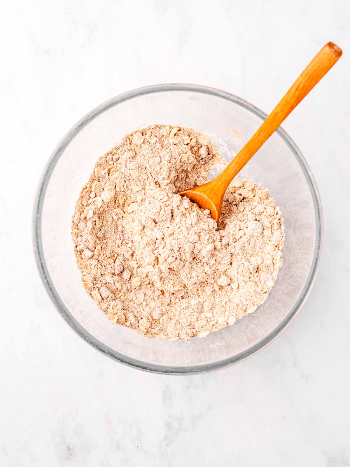 overhead view of dry ingredients for crumble topping in glass bowl with wooden spoon