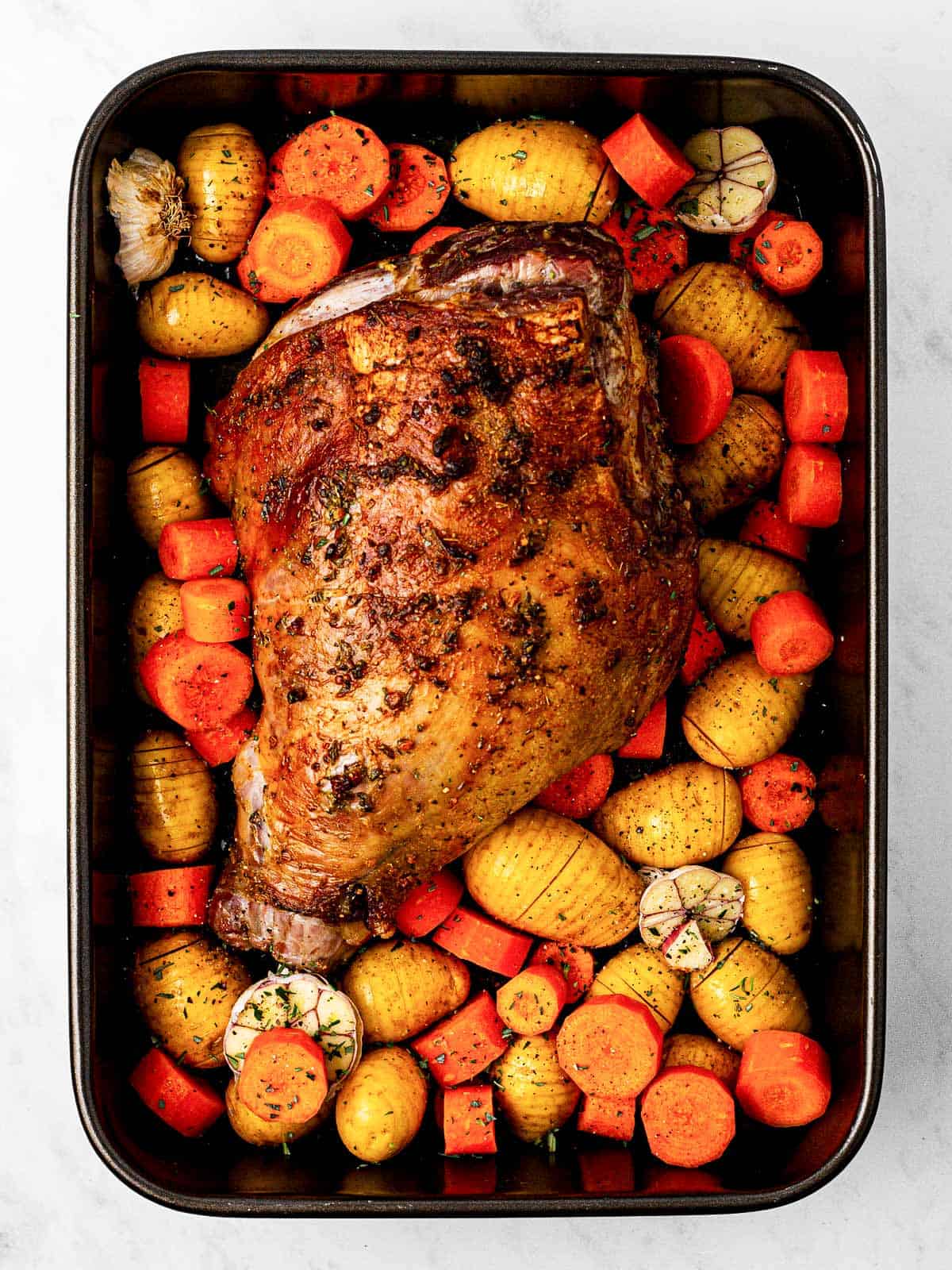 overhead view of roasted leg of lamb with raw carrots and baby potatoes in black roasting pan