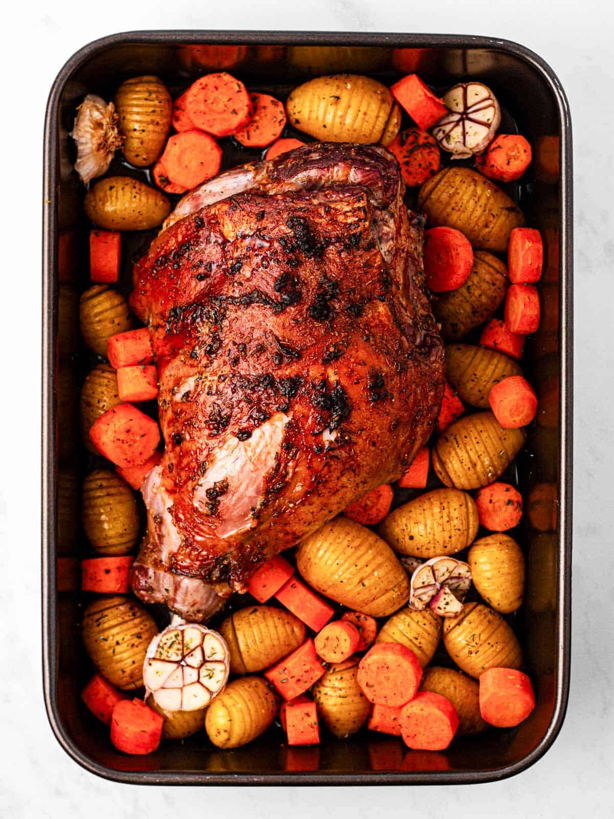 overhead view of roasted leg of lamb with roasted carrots and baby potatoes in black roasting pan