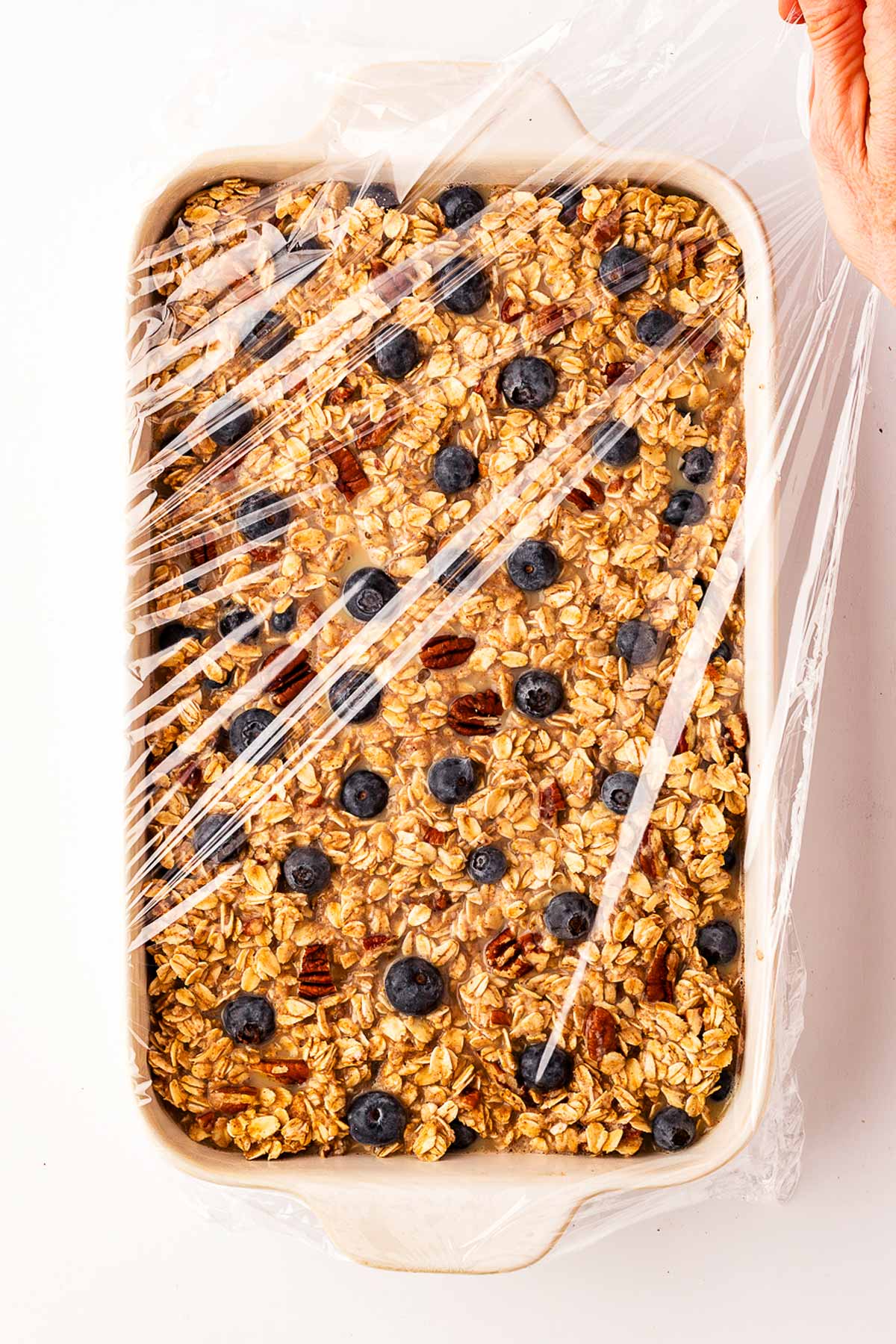 overhead view of female hand covering unbaked blueberry oatmeal in casserole dish with plastic wrap