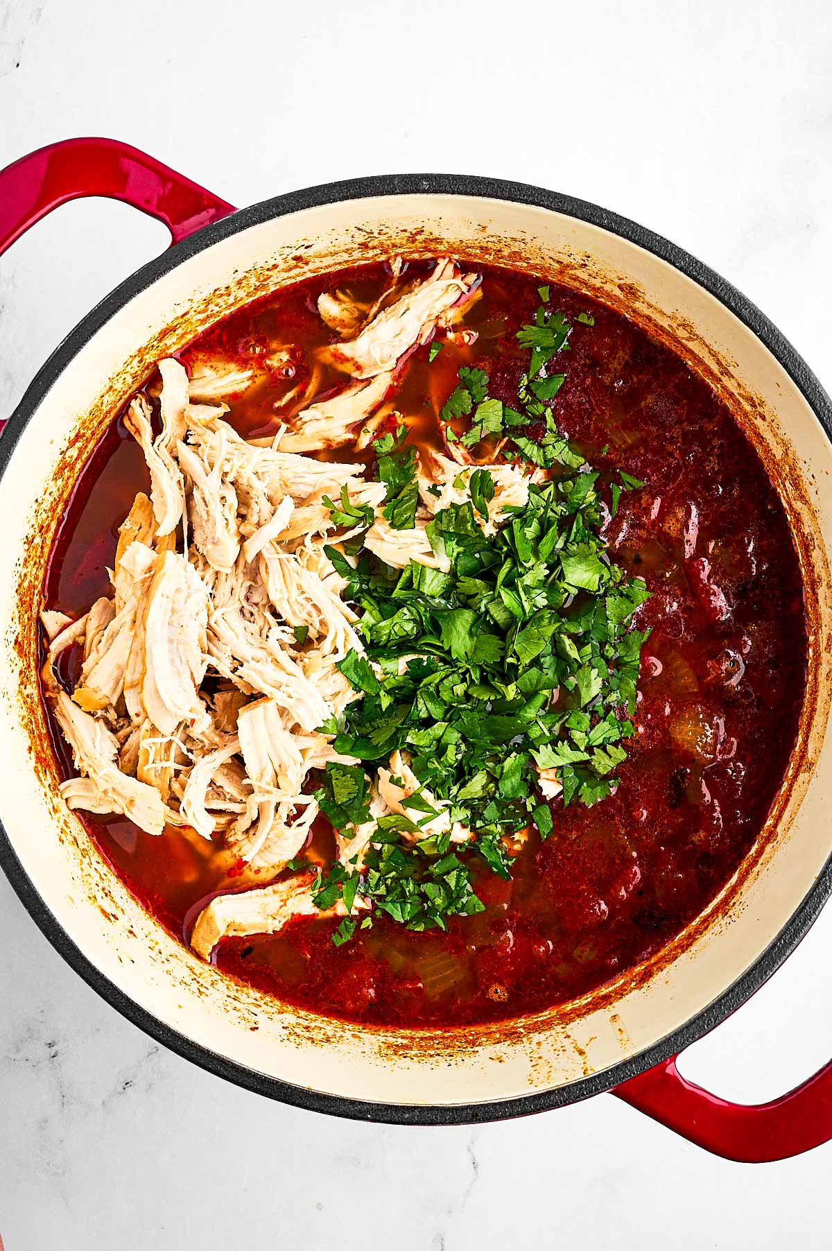 overhead view of shredded chicken and chopped cilantro on top of tomato broth in pot