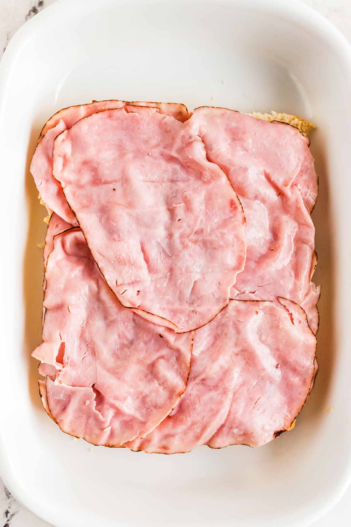 overhead view of deli ham on halved slab of dinner rolls in white baking dish