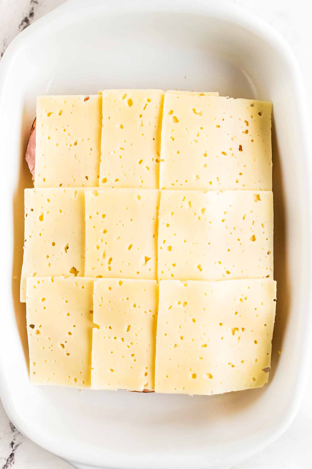 overhead view of cheese slices and deli ham on halved slab of dinner rolls in white baking dish