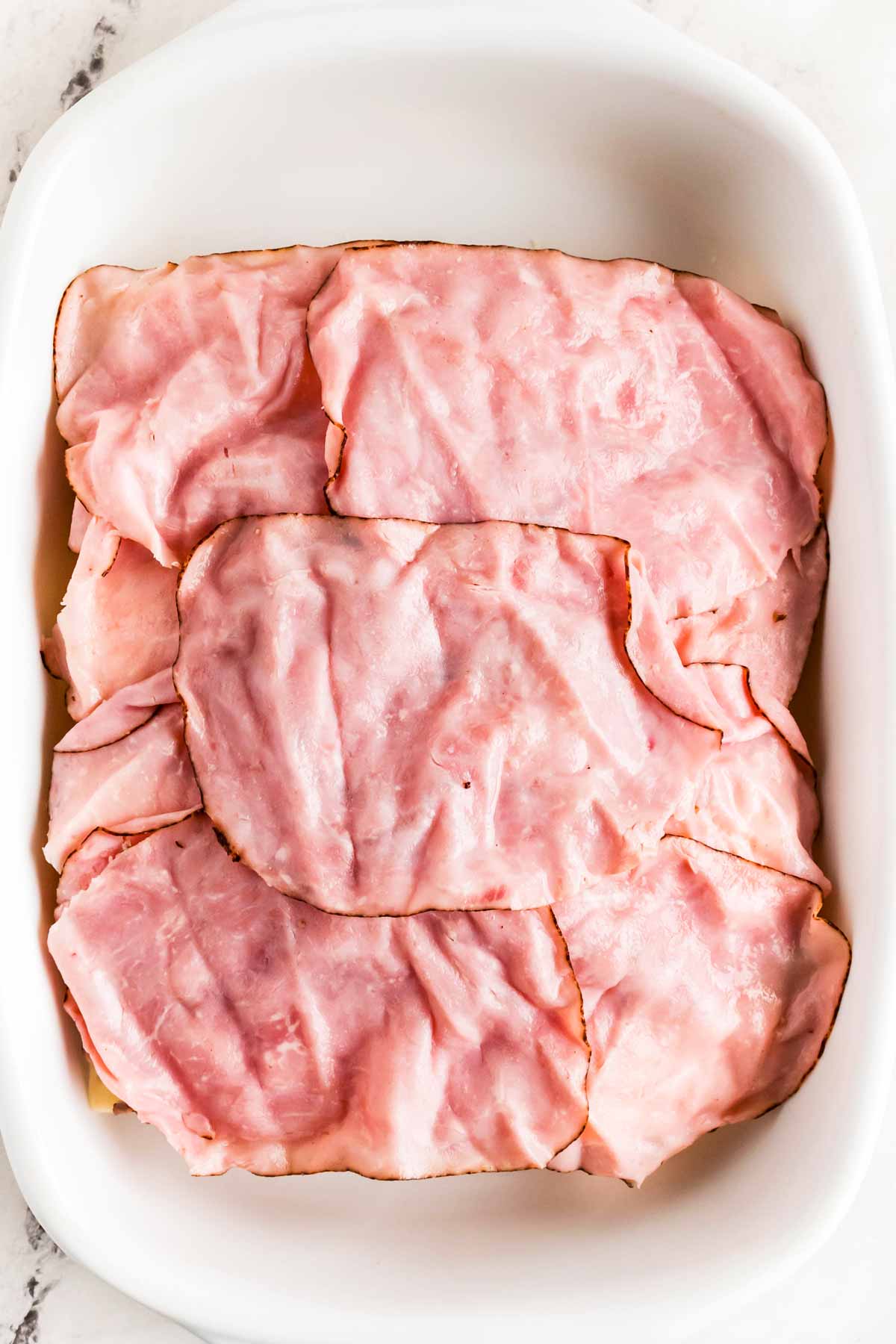 overhead view of deli ham on top of cheese on halved slab of dinner rolls in white baking dish