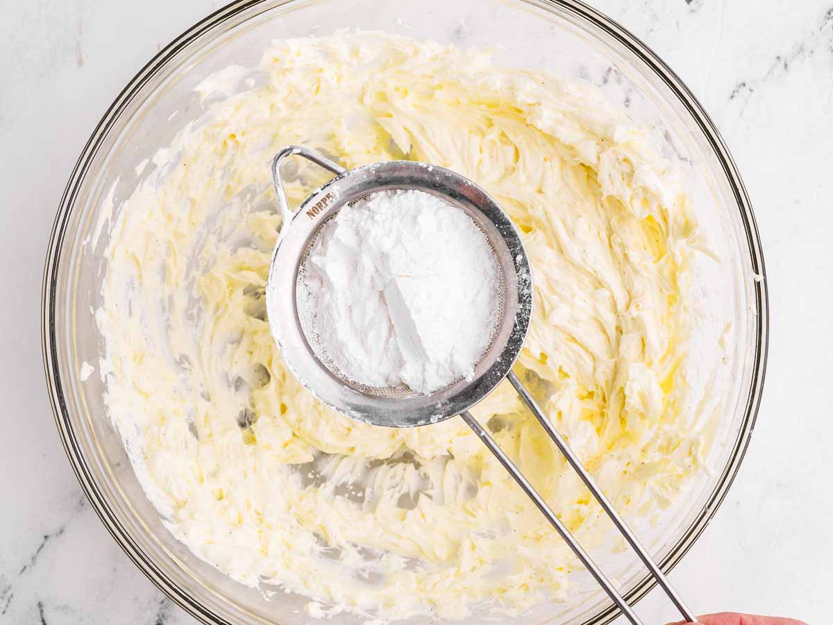 overhead view of icing sugar in small sieve over bowl with beaten cream cheese