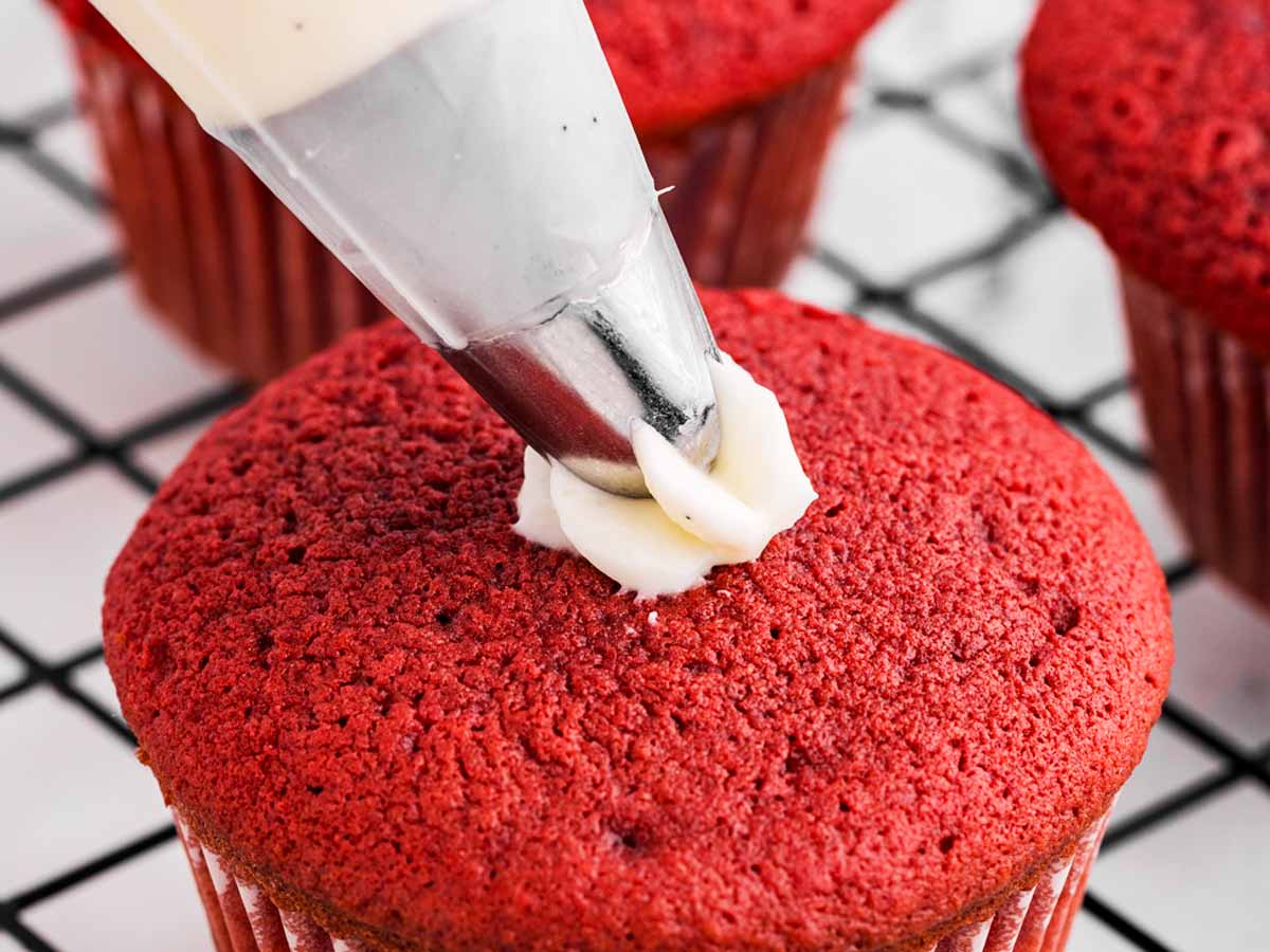 overhead view of piping bag piping cream cheese frosting onto red velvet cupcake