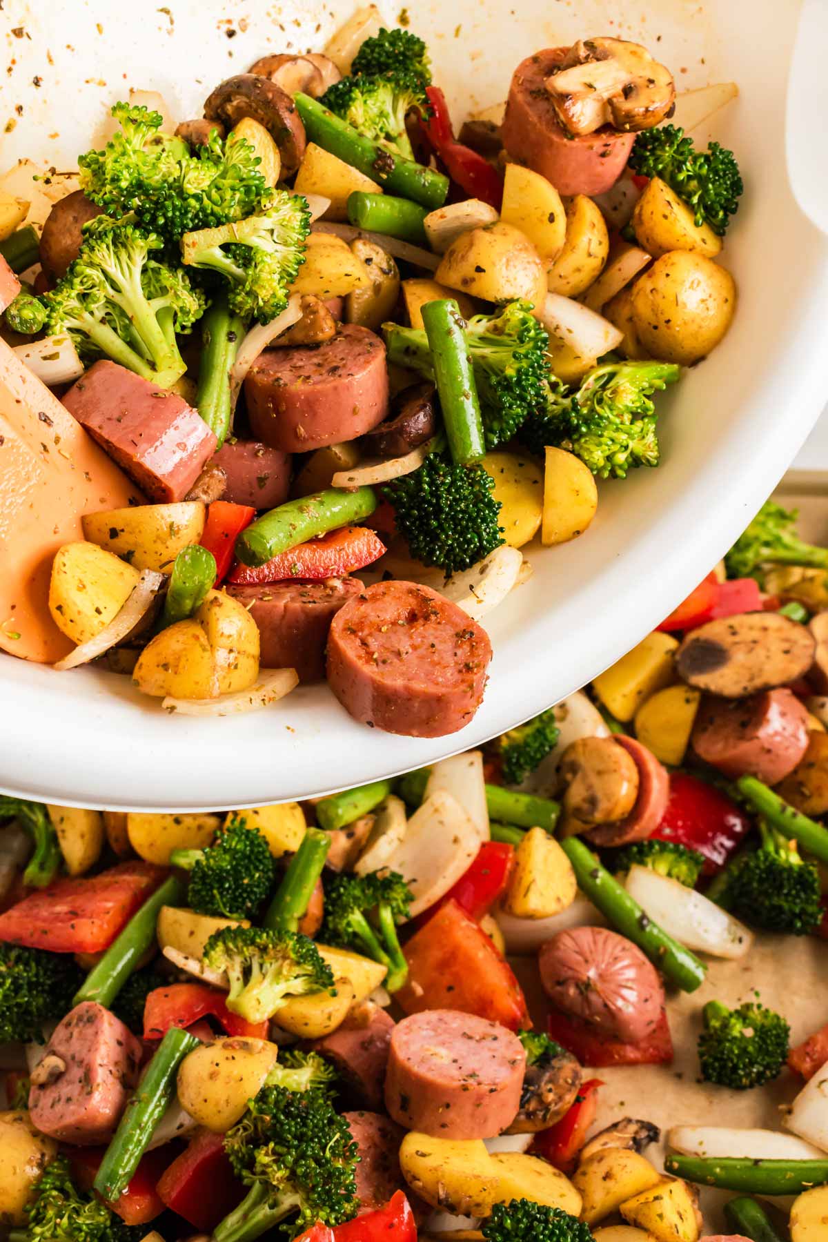 frontal view of sausage and vegetables being transferred from bowl to lined sheet pan