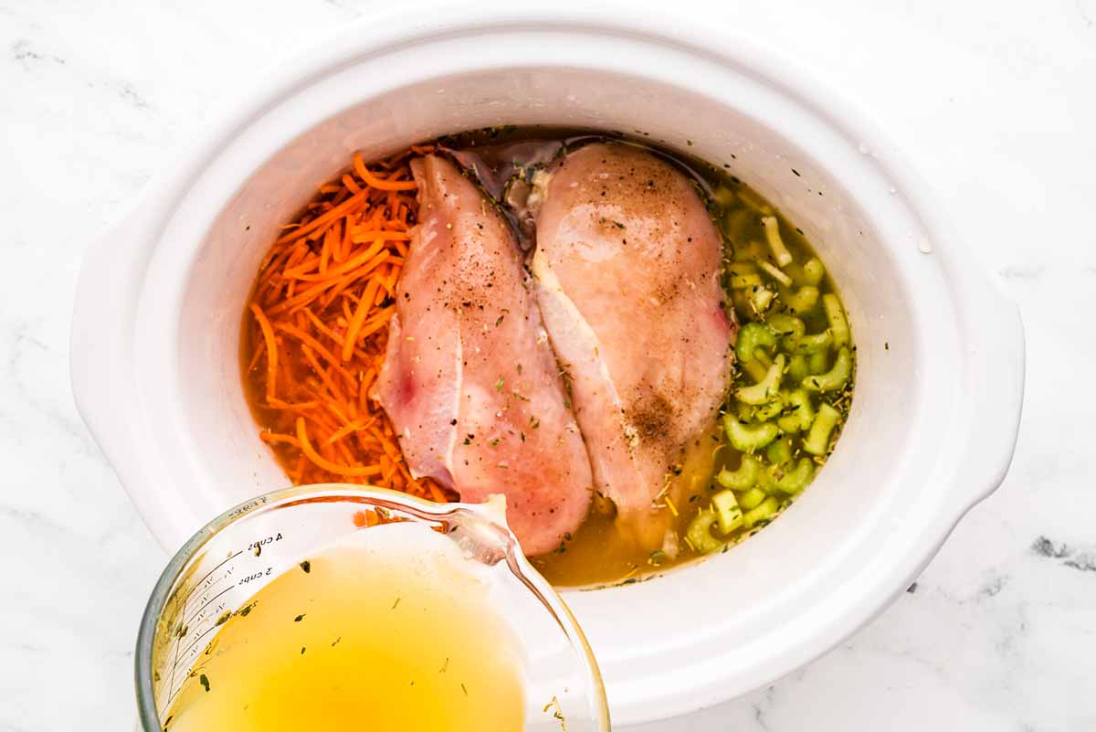 overhead view of chicken breast and vegetables in slow cooker with chicken broth being poured over