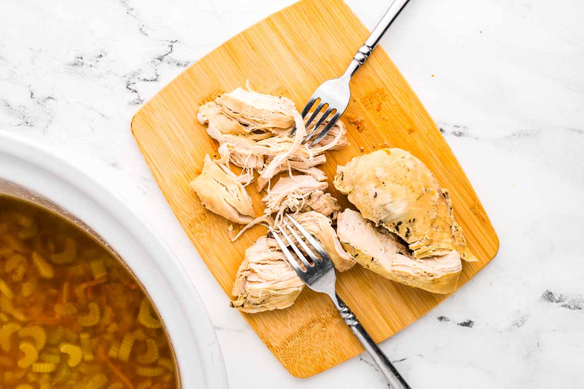 overhead view of shredded chicken on wooden board with two forks next to slow cooker