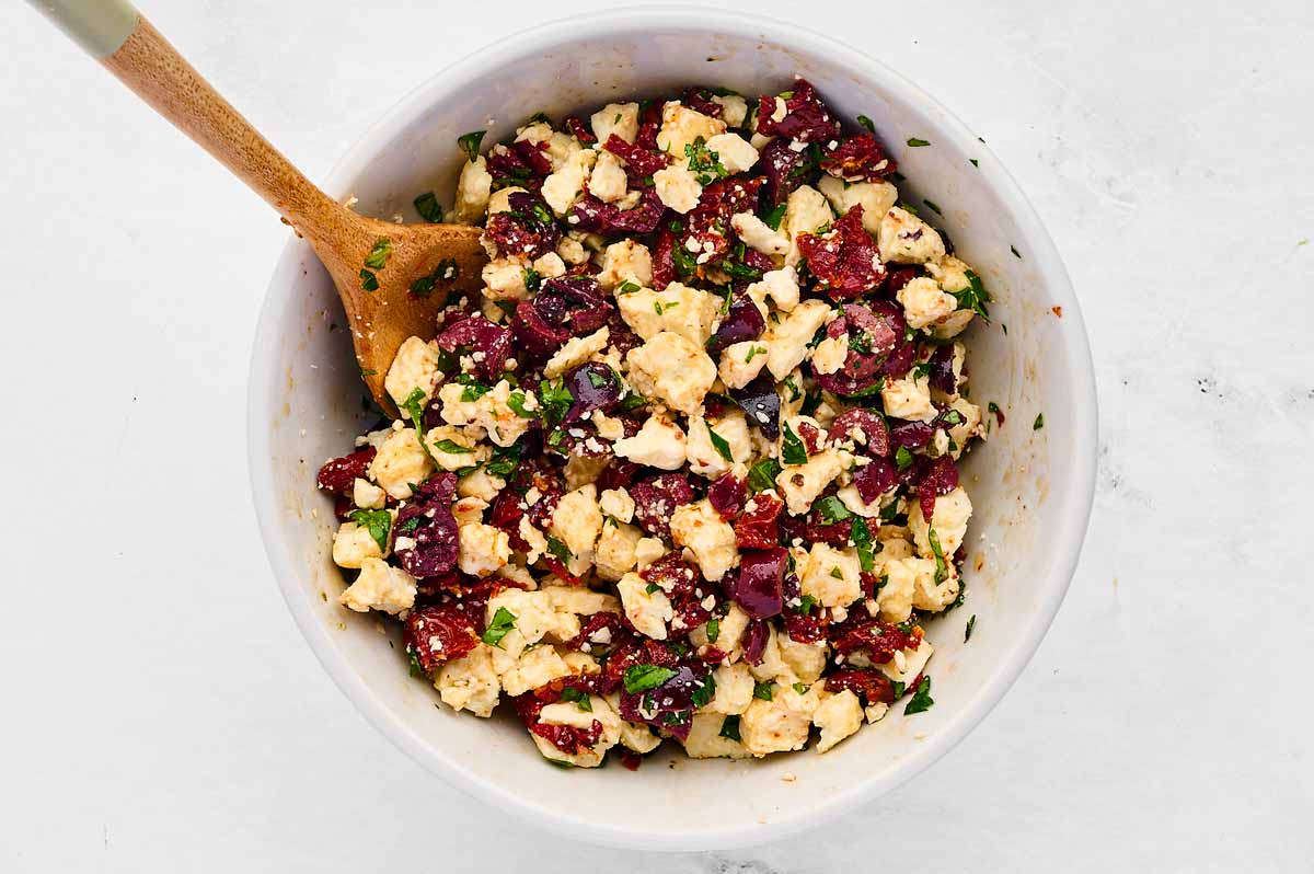 overhead view of feta cheese, olives and herbs in white bowl with wooden spoon