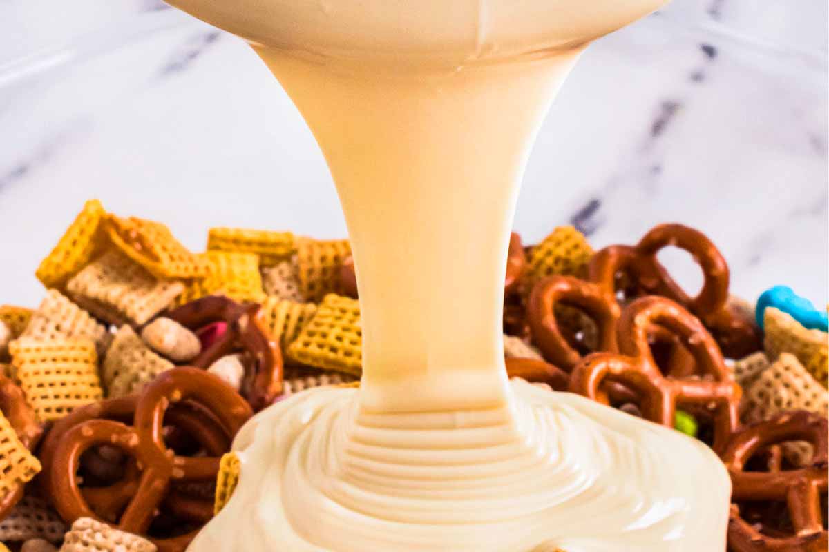 frontal close up view of melted white chocolate pouring over pretzels and Chex cereal in bowl